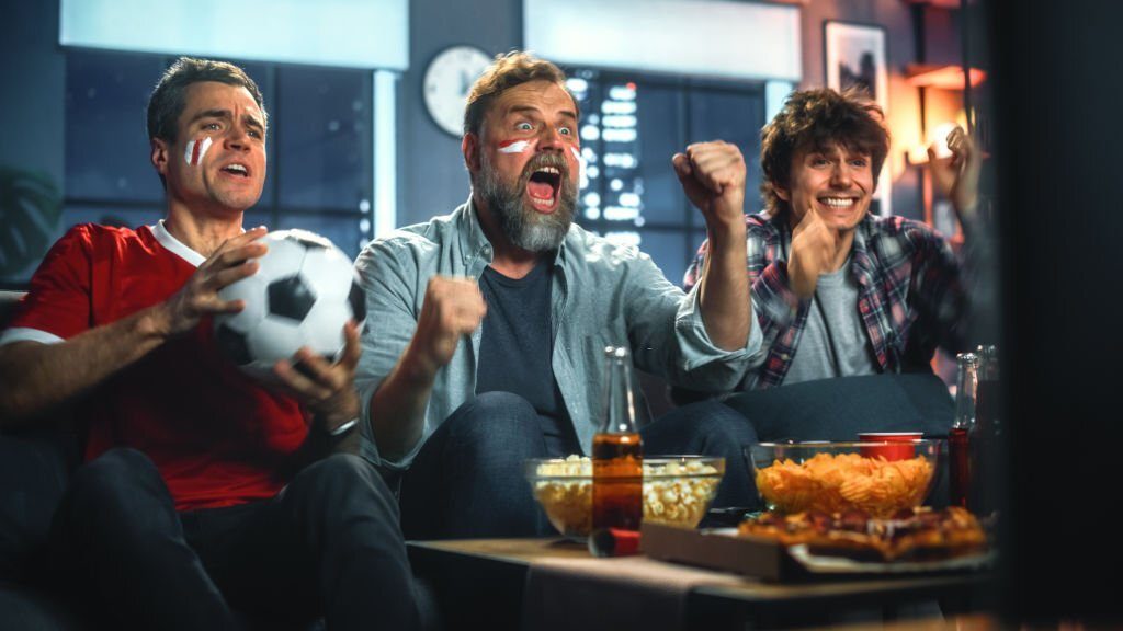 At Home Three Joyfuls Sports Fans with Painted Faces Sitting on a Couch Watch Game on TV, Celebrate Victory when Sports Team Wins Championship. Friends Cheer, Shout.